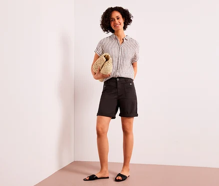 Une femme souriante aux cheveux bouclés se tient debout, tenant une pochette tressée. Elle porte une chemise rayée, un bermuda Street One « Yulius » marron et des sandales noires.