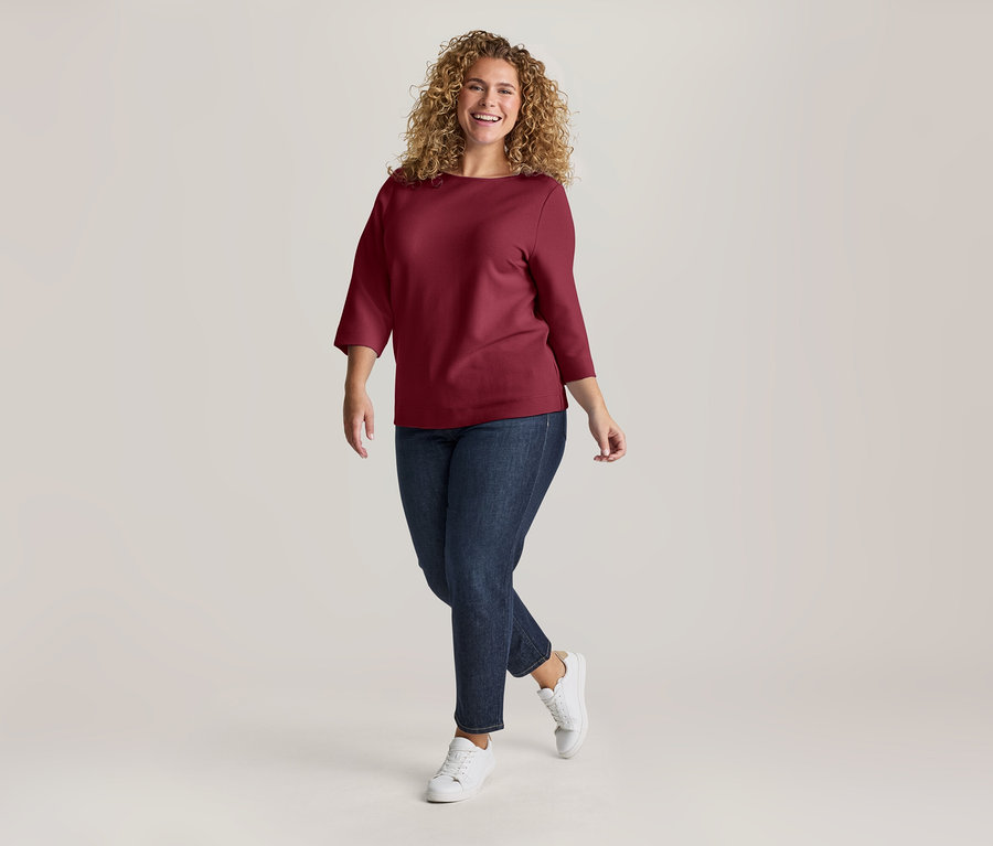 Femme souriante aux cheveux bouclés portant un haut bordeaux, un jean bleu foncé et des baskets blanches, marchant vers l'avant.