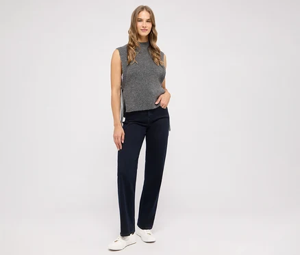 Femme debout portant un haut gris sans manches, un jean bleu foncé et des baskets blanches, la main gauche dans la poche.