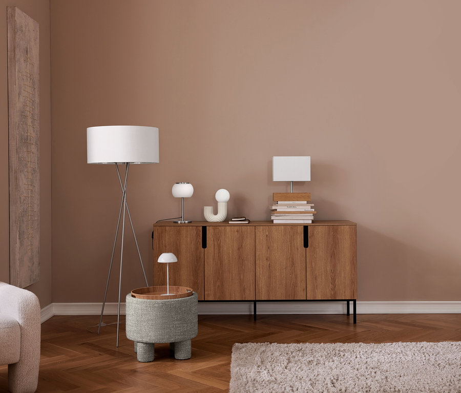 Un salon moderne avec un fauteuil beige clair, un parquet en chevrons et un mur rose poudré. Un lampadaire trépied argenté se tient à côté d'un ottoman gris texturé avec un plateau en bois et une petite lampe blanche. Un buffet en bois abrite deux lampes de table blanches, des objets décoratifs et des piles de livres. Un tapis shaggy clair est au sol.
