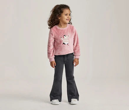 Jeune fille aux cheveux bouclés debout, portant un pull rose duveteux avec un motif de pingouin et un jean évasé gris foncé.