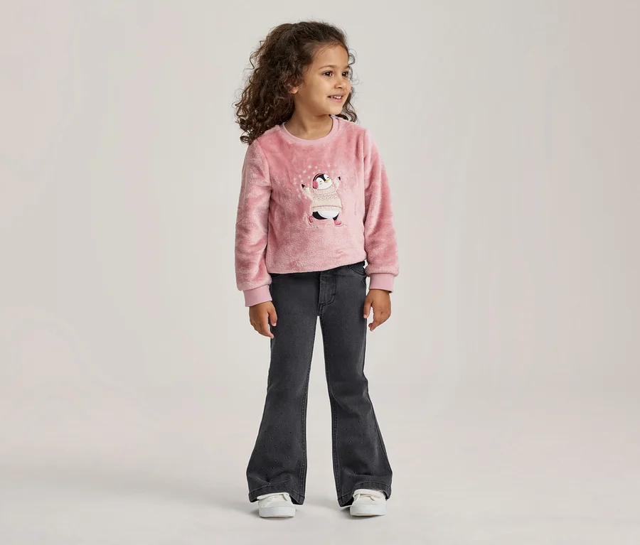 Jeune fille aux cheveux bouclés debout, portant un pull rose duveteux avec un motif de pingouin et un jean évasé gris foncé.