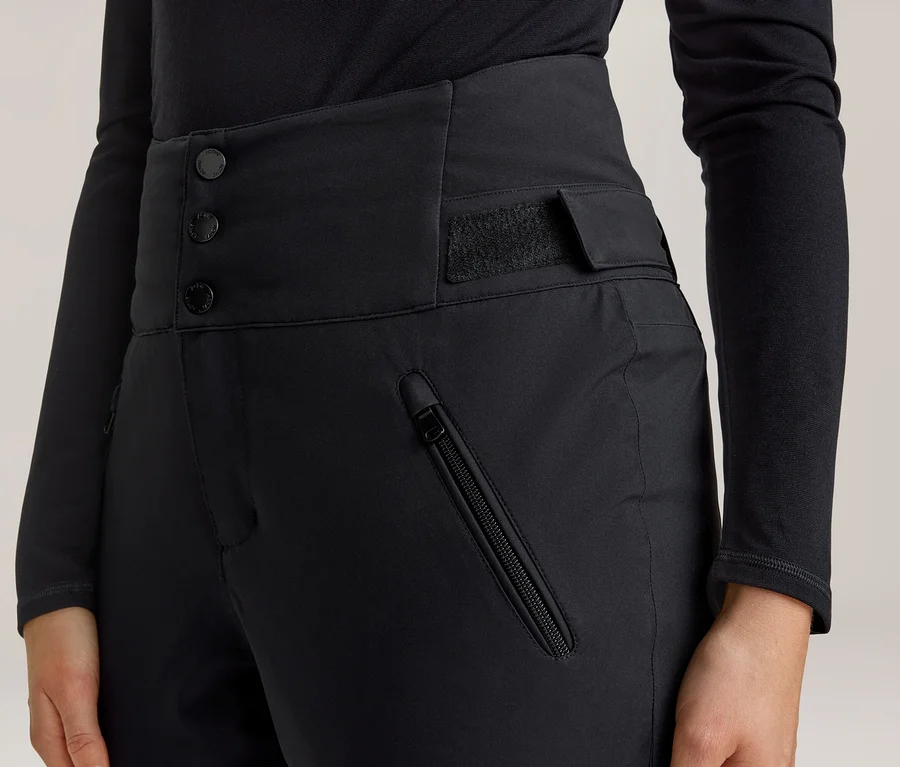 Gros plan d'une personne portant un pantalon noir taille haute avec trois boutons et une bande velcro à la taille, ainsi qu'une poche zippée sur la cuisse droite. La personne porte également un haut noir à manches longues.