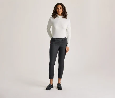 Une femme aux cheveux bouclés foncés est debout, vêtue d'un col roulé blanc à manches longues et d'un tregging gris foncé – coupe « Nelly », les mains dans les poches.