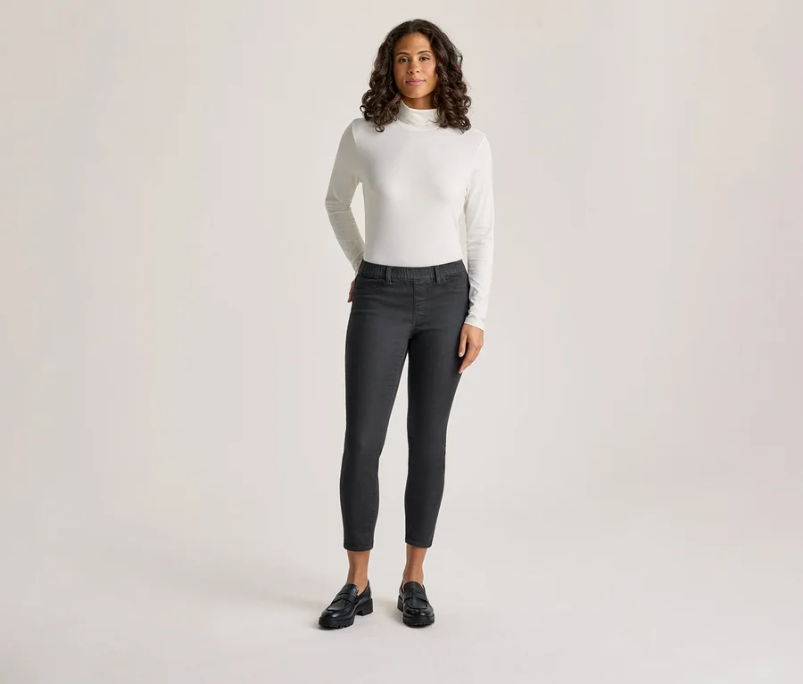 Une femme aux cheveux bouclés foncés est debout, vêtue d'un col roulé blanc à manches longues et d'un tregging gris foncé – coupe « Nelly », les mains dans les poches.