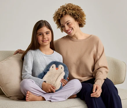 Une femme adulte et une jeune fille sont assises sur un canapé, toutes deux souriantes. La fille, à gauche, tient une peluche pingouin bleue et beige. La femme, à droite, a son bras autour de la fille.