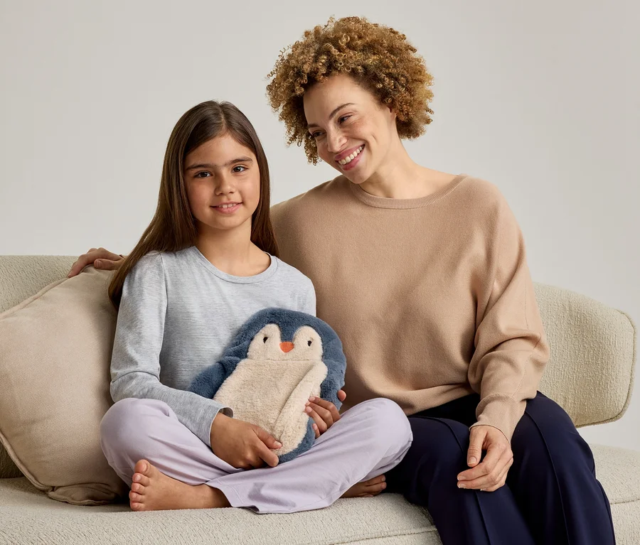 Une femme adulte et une jeune fille sont assises sur un canapé, toutes deux souriantes. La fille, à gauche, tient une peluche pingouin bleue et beige. La femme, à droite, a son bras autour de la fille.
