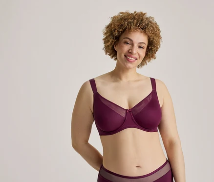 Une femme souriante aux cheveux bouclés et aux taches de rousseur porte un soutien-gorge couleur prune avec des détails en maille et une culotte assortie, les mains derrière le dos.
