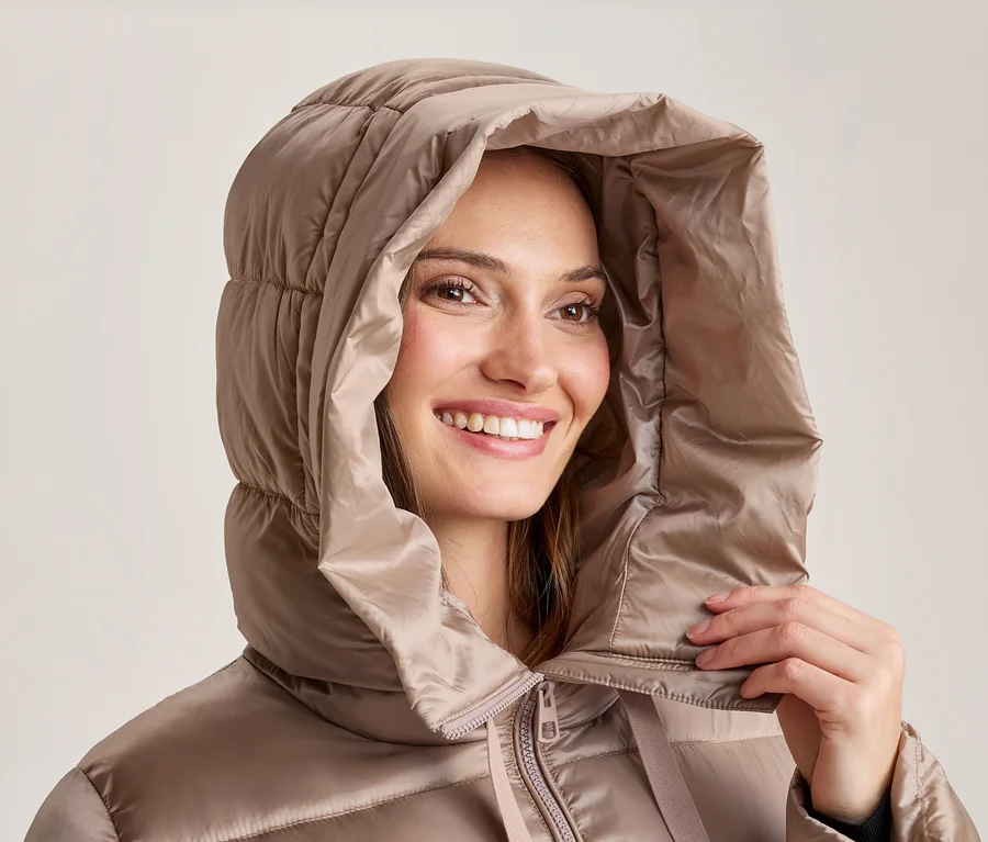 Femme souriante portant une doudoune beige avec la capuche relevée, une main près du col.