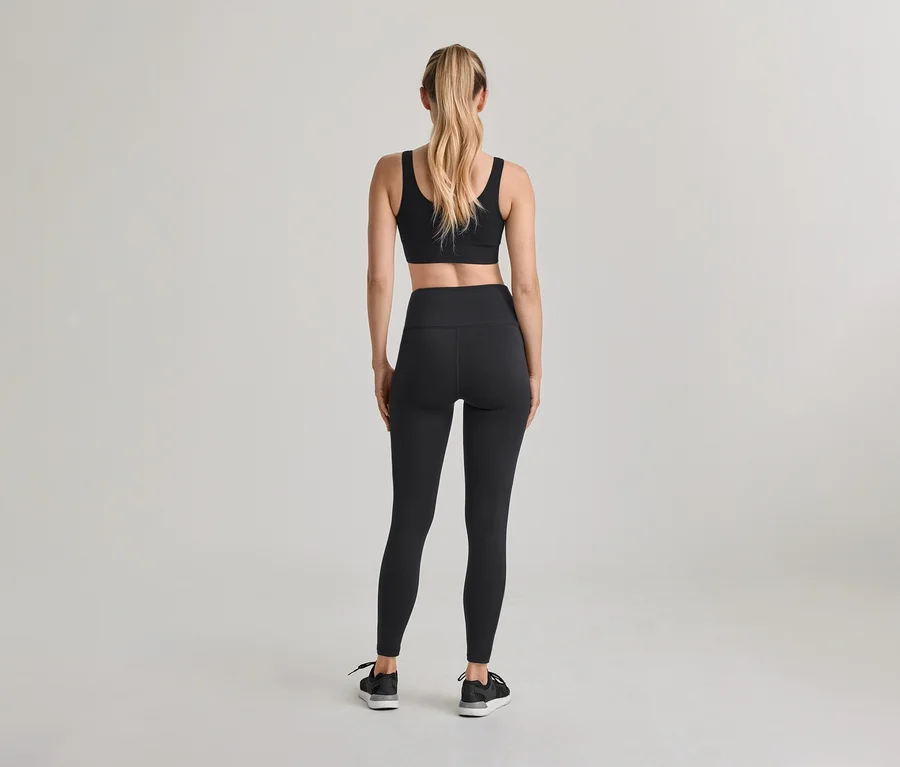 Femme de dos, portant un soutien-gorge de sport noir, un legging noir taille haute et des baskets noires. Ses cheveux blonds sont attachés en queue de cheval.