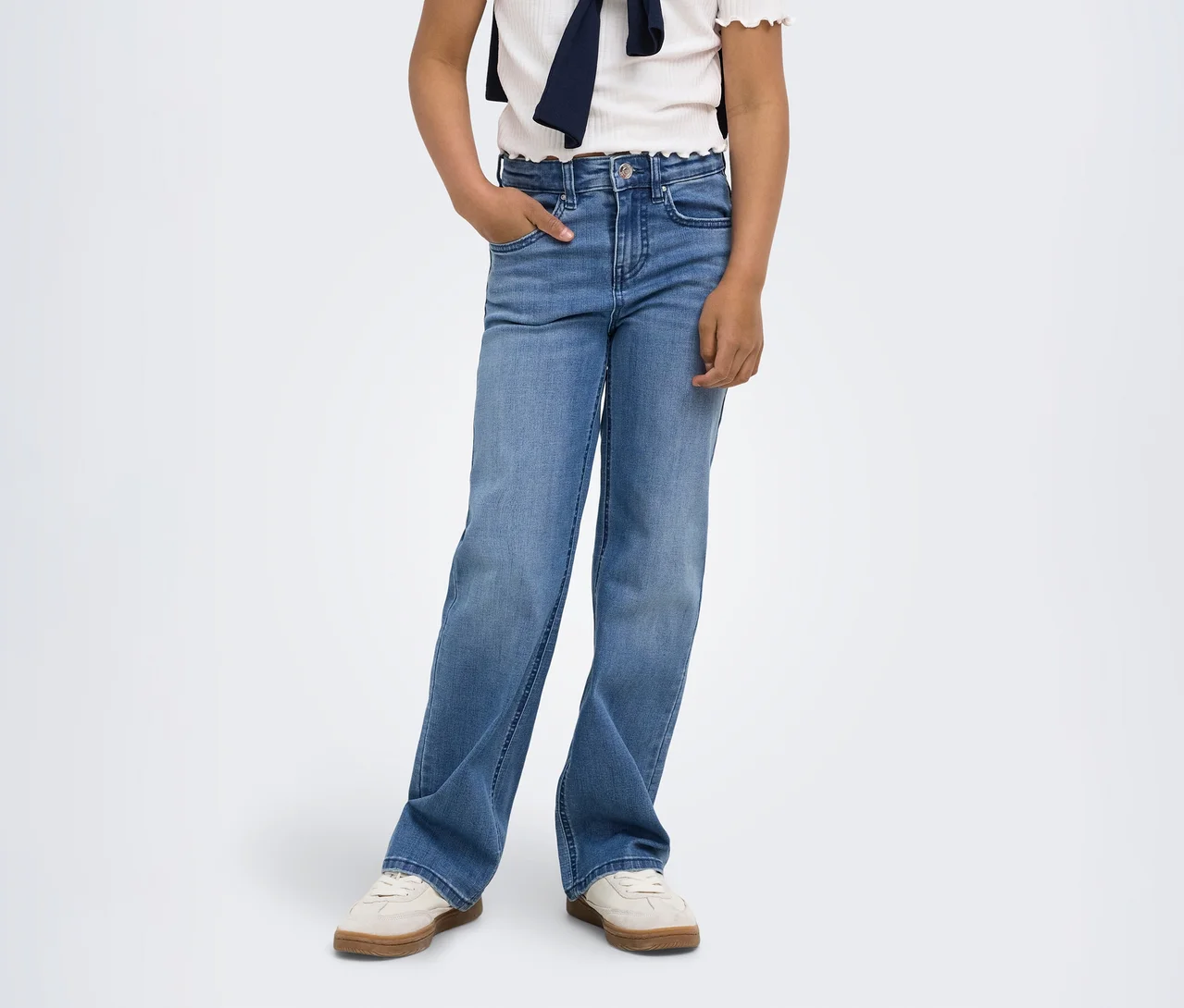 Une personne porte un jean bleu clair ample, un haut blanc et un pull bleu marine noué autour des épaules. Une main est dans la poche, avec des baskets claires à semelles marron.