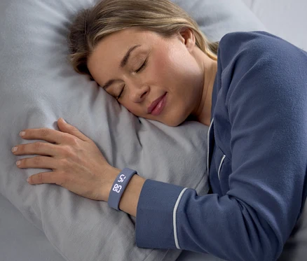 Une femme dort paisiblement sur le côté sur un oreiller gris, vêtue d'un pyjama bleu. Un bracelet connecté gris à son poignet gauche affiche "58" et "80".