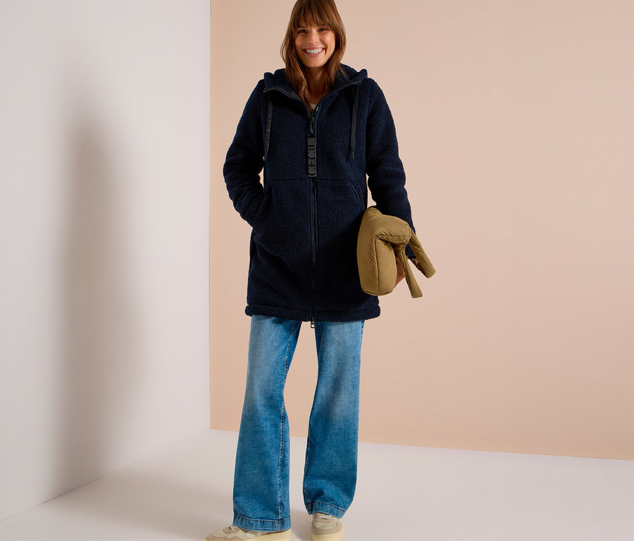 Femme souriante portant une veste CECIL bleu marine à capuche et en tissu pelucheux, un jean large bleu clair et des baskets beiges, tenant une doudoune beige pliée.