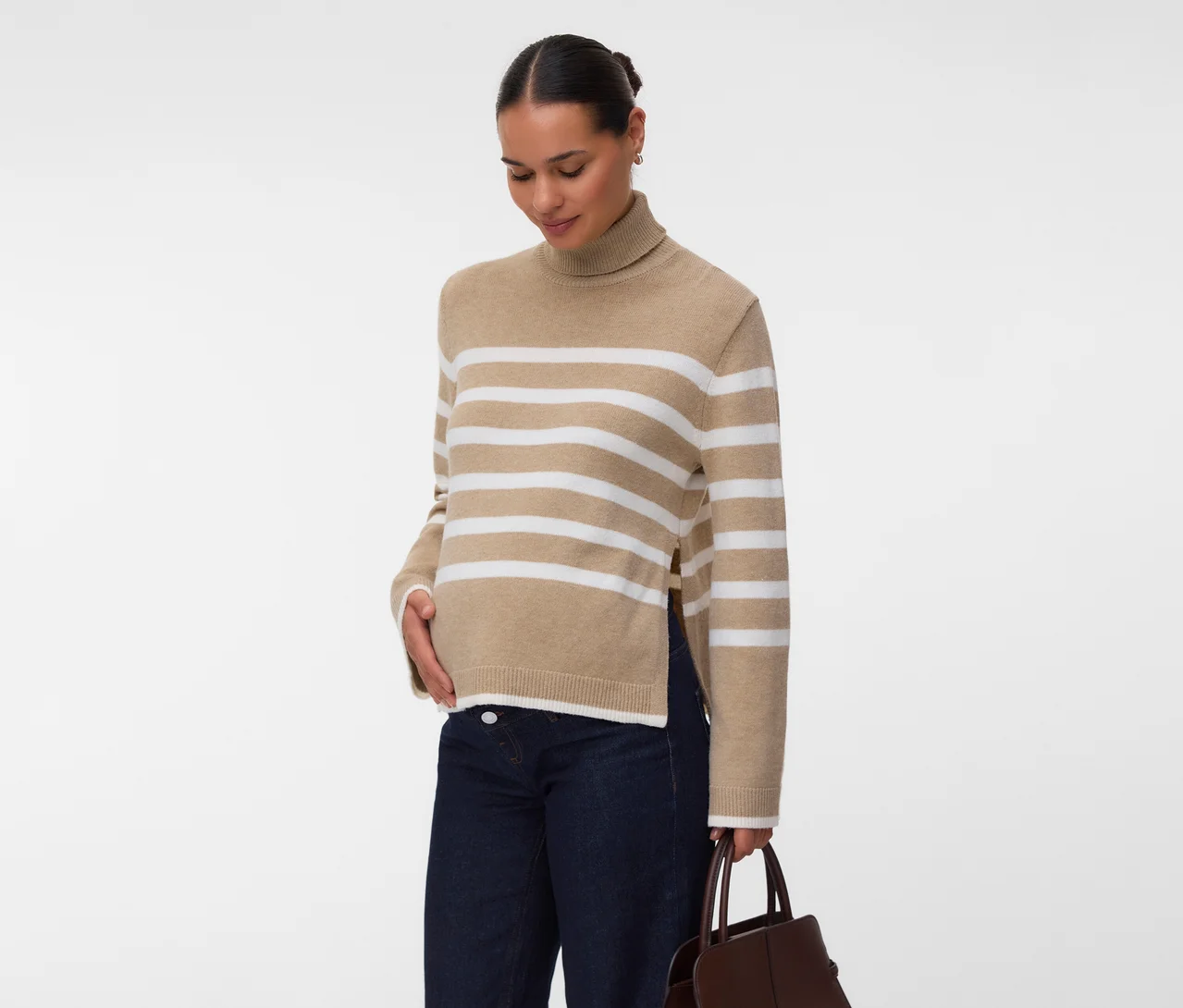 Femme portant un pull à col roulé beige à rayures blanches avec fentes latérales, un jean foncé et tenant un sac à main marron, la main sur le ventre.