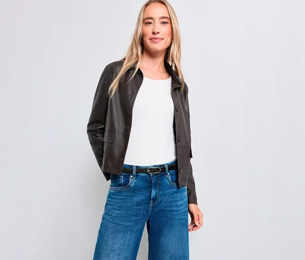 Une femme blonde se tient debout, souriant légèrement, devant un fond blanc. Elle porte une veste marron foncé, un haut blanc, un jean bleu et une ceinture foncée.