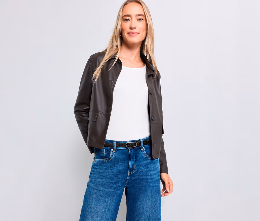 Une femme blonde se tient debout, souriant légèrement, devant un fond blanc. Elle porte une veste marron foncé, un haut blanc, un jean bleu et une ceinture foncée.