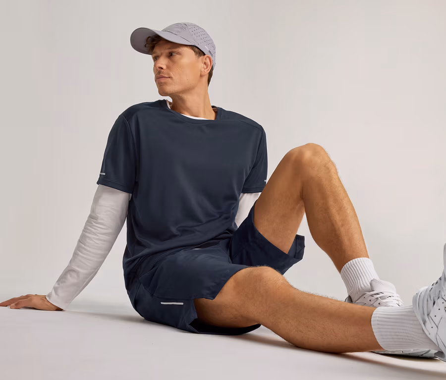 Un homme est assis par terre, portant une casquette grise, un t-shirt marine sur un t-shirt blanc à manches longues, un short de sport bleu marine, des chaussettes blanches et des baskets blanches, regardant vers la droite.