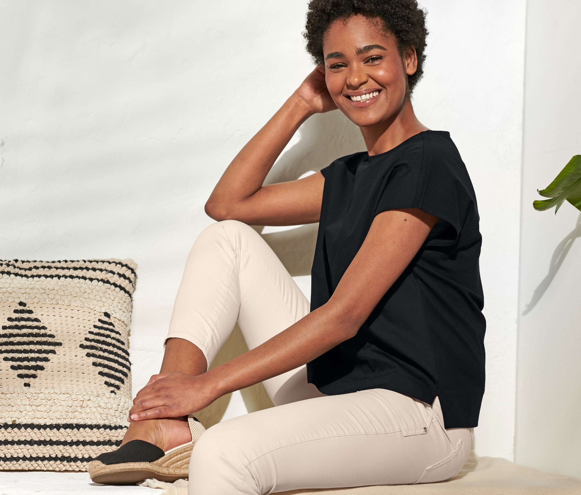 Une femme souriante est assise, portant une blouse bimatière noire et un pantalon clair.