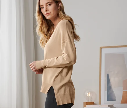 Une femme blonde pose avec un pull en maille fine beige et un pantalon noir.