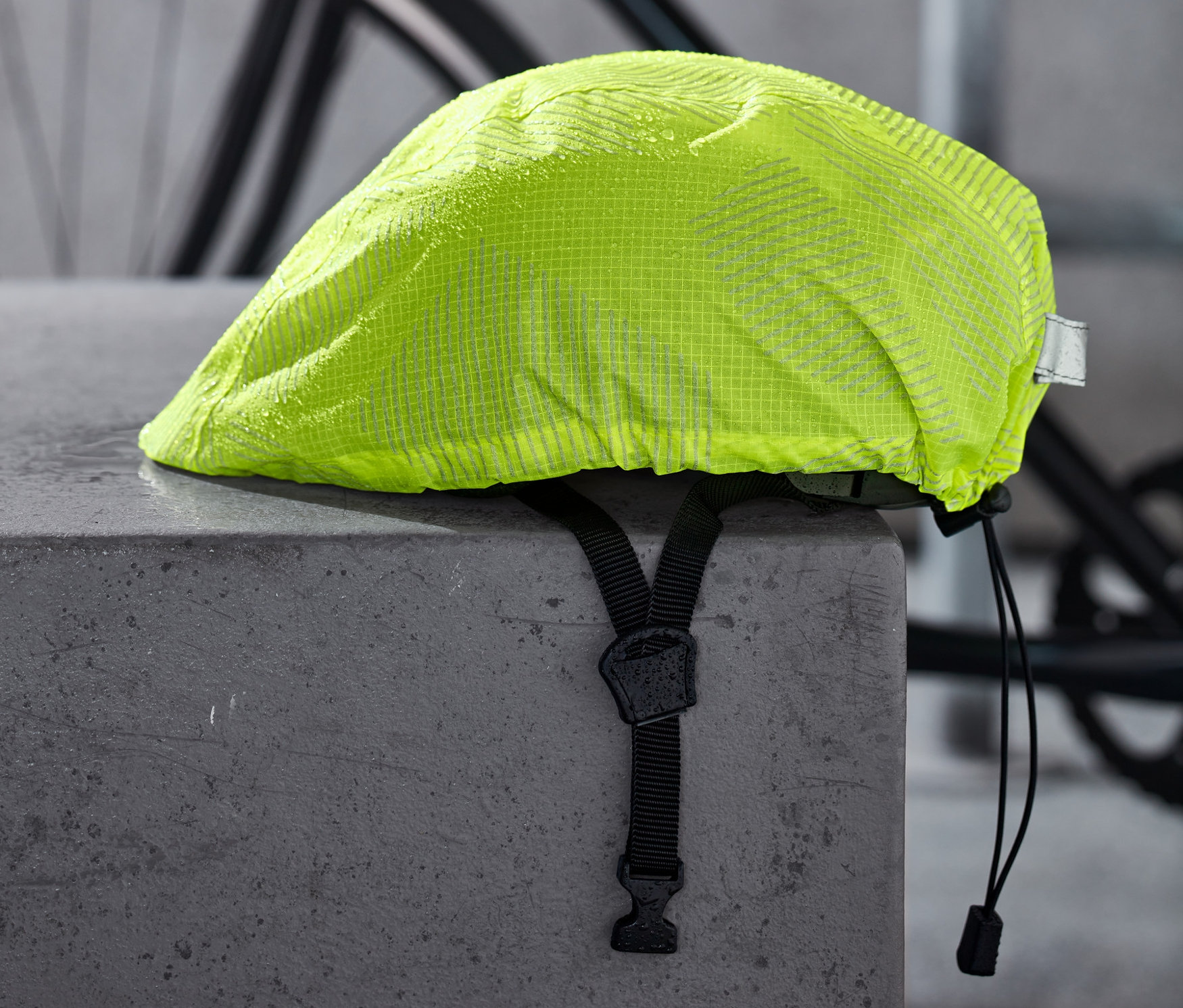 Une housse de pluie jaune néon pour casque de vélo repose sur un bloc de béton gris, avec un vélo en arrière-plan.