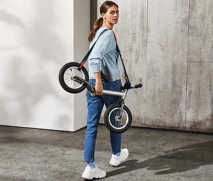 Femme portant un vélo d'équilibre argenté sur l'épaule avec une bandoulière multifonction.