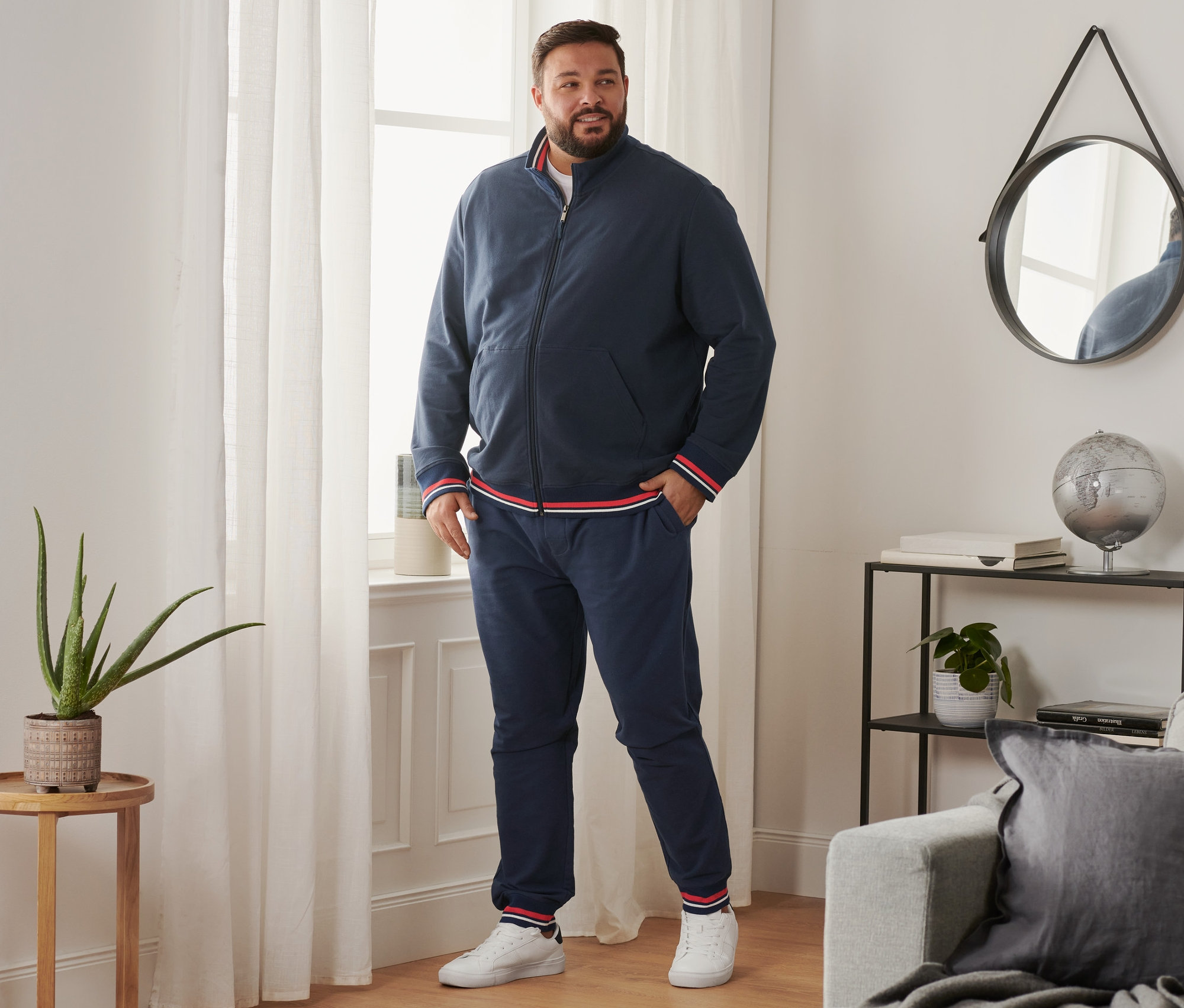 Un homme se tient près de la fenêtre et porte un pantalon de jogging, des tennis et une veste molletonnée.