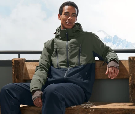 Un homme est assis sur un banc et porte une veste de ski kaki et des chaussures de ski.