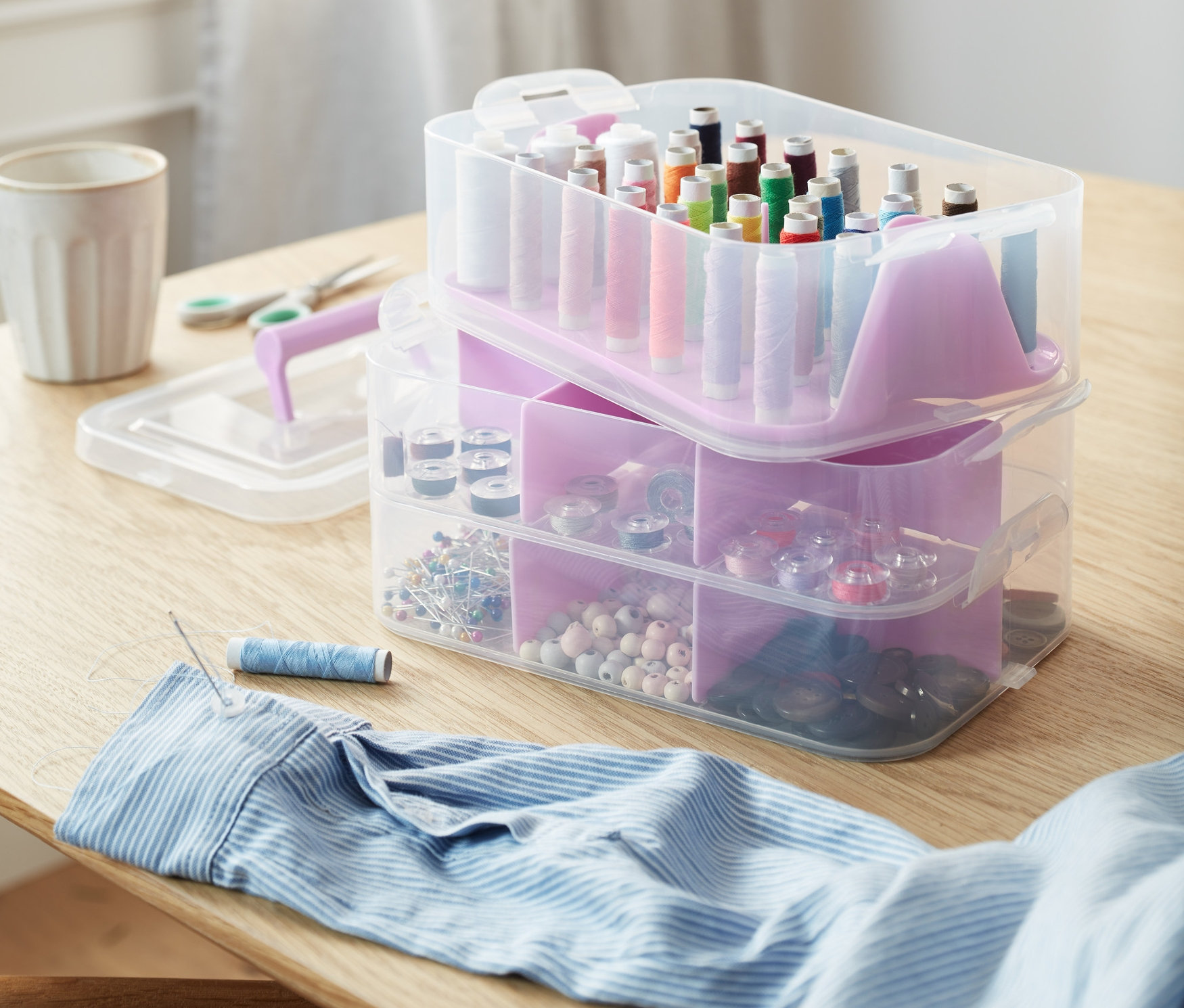 Sur la table, il y a des boîtes de rangement, des ciseaux, des boutons, des bobines de fil et une aiguille avec du fil dans une chemise.
