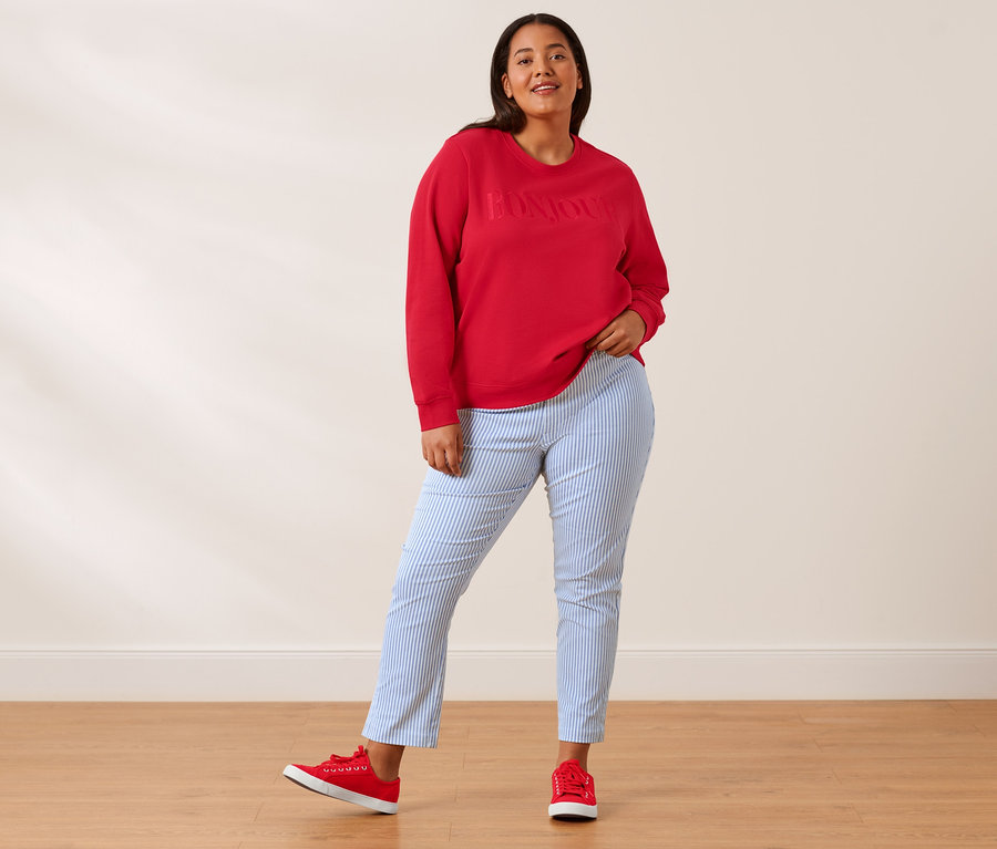 Femme posant dans un sweat-shirt rouge avec broderie, un pantalon stretch à rayures et des baskets en toile.