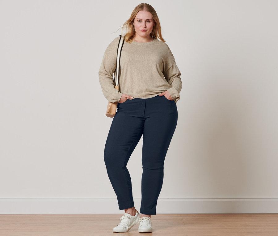 Femme posant dans un pull en tricot fin beige avec détail nervuré, un pantalon en bengaline bleu foncé, des baskets en cuir et un sac à bandoulière.
