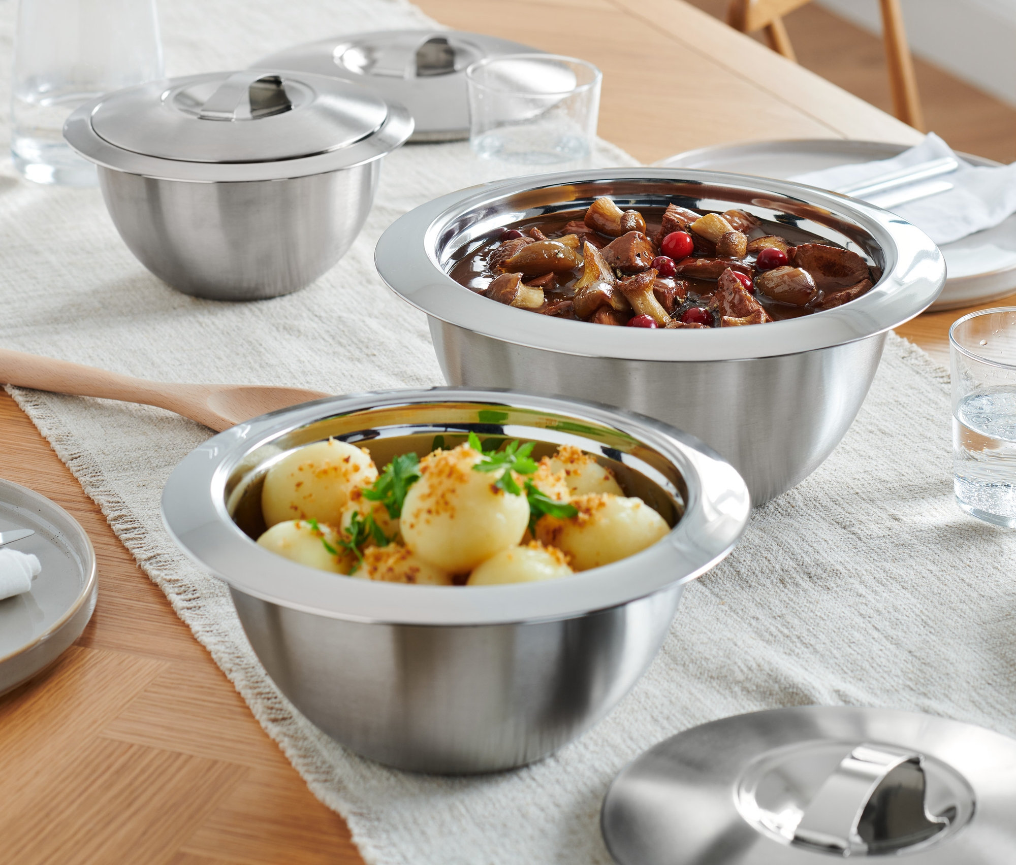 Sur une table en bois, il y a trois saladiers thermiques de service avec couvercles en inox, des assiettes et des verres d'eau.