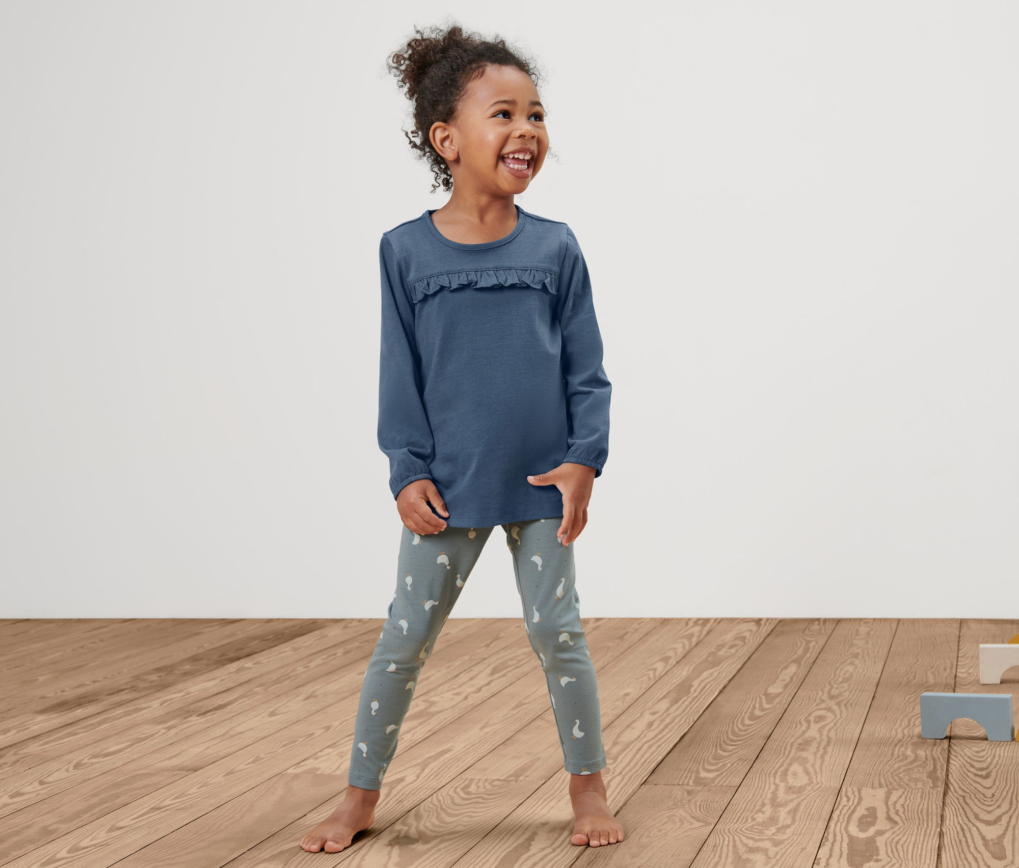 Une petite fille souriante se tient sur un plancher en bois et porte un t-shirt pour tout-petit et des leggings.