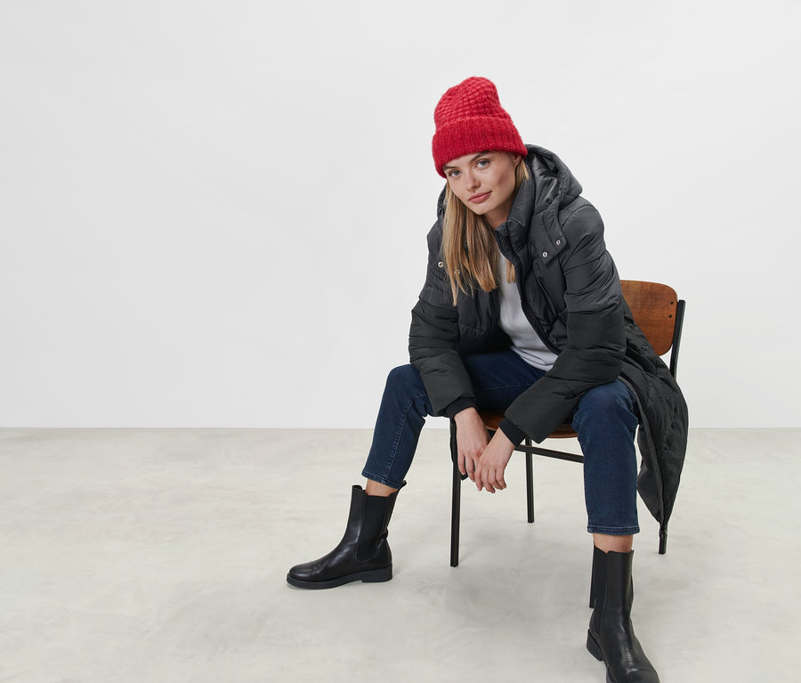 Une femme est assise sur une chaise et porte un manteau matelassé à capuche et un bonnet rouge.