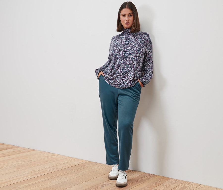 Femme posant en blouse à col montant, mille-fleurs, pantalon de jogging et baskets en cuir.