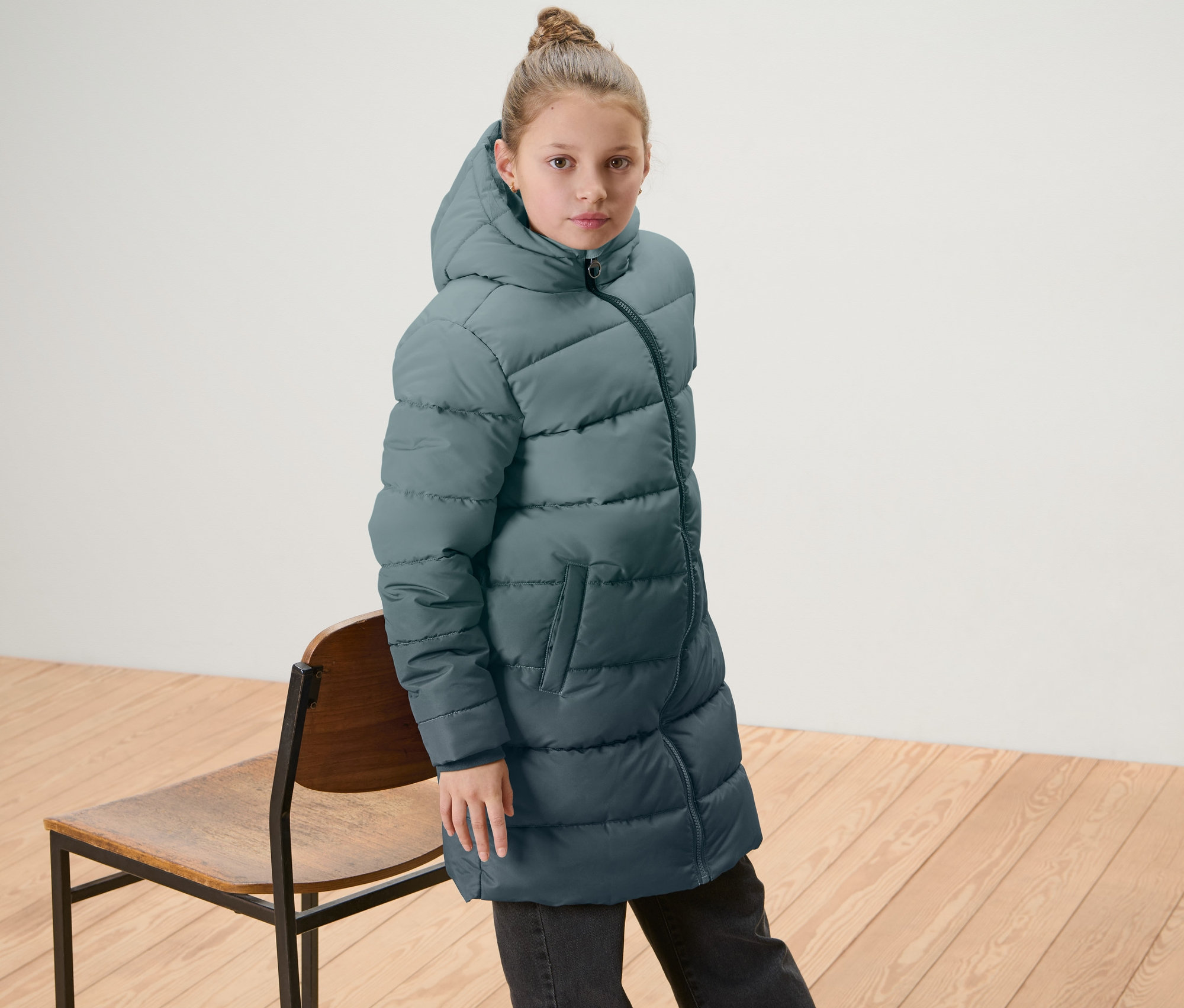 Une fille porte un manteau thermique pour enfant aux couleurs dégradées et se tient à côté d'une chaise en bois.