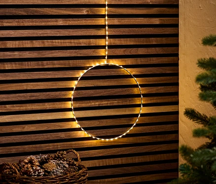 Un cercle à LED d'extérieur suspendu devant un mur en bois, avec un panier de pommes de pin en dessous.