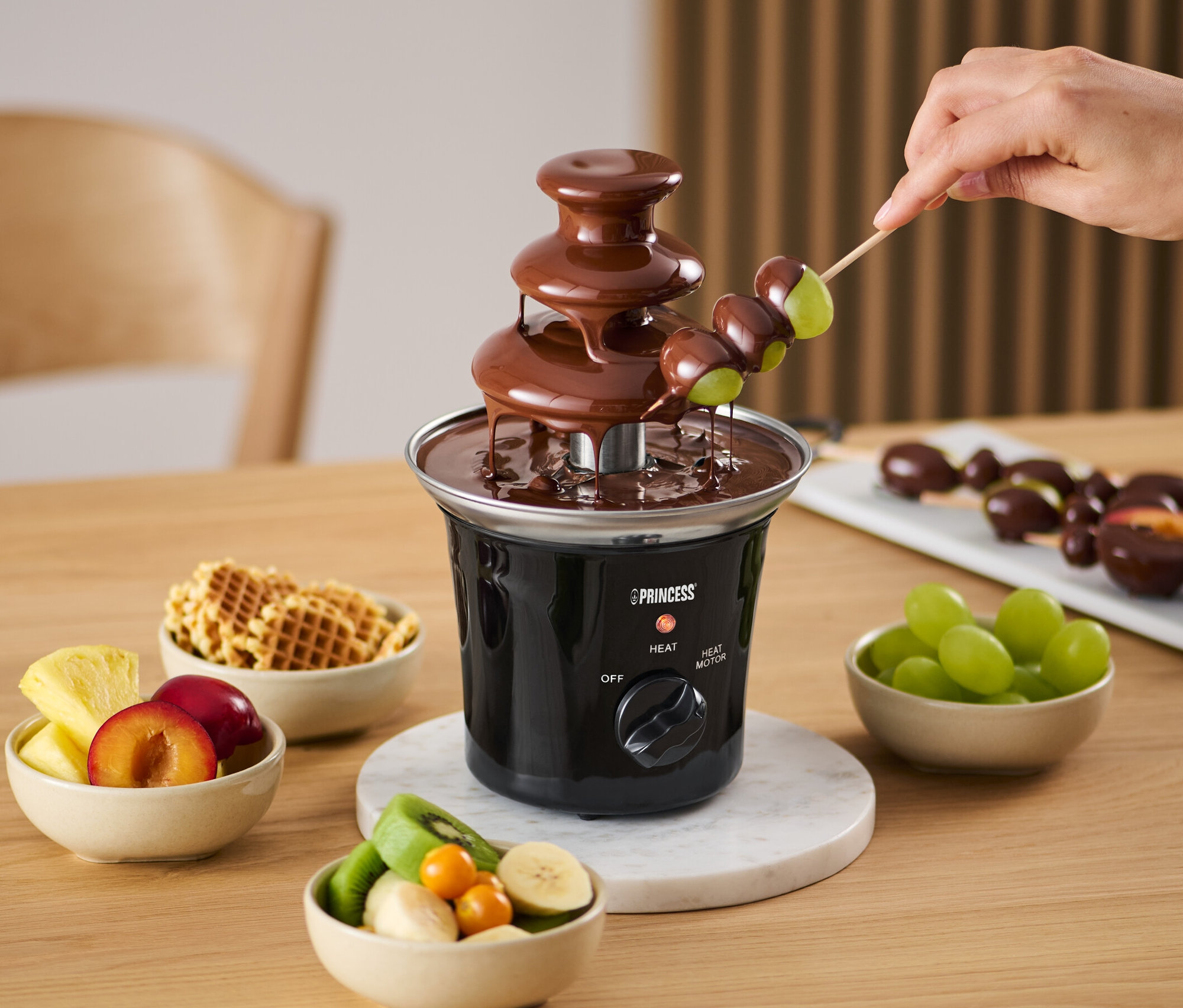 Une main trempe des raisins sur une brochette dans une mini-fontaine à chocolat électrique. Des coupelles de fruits et des gaufres sont également disponibles.