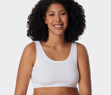 Femme souriante aux cheveux bouclés portant une brassière SCHIESSER blanche et une culotte blanche, appuyée sur un bloc blanc.
