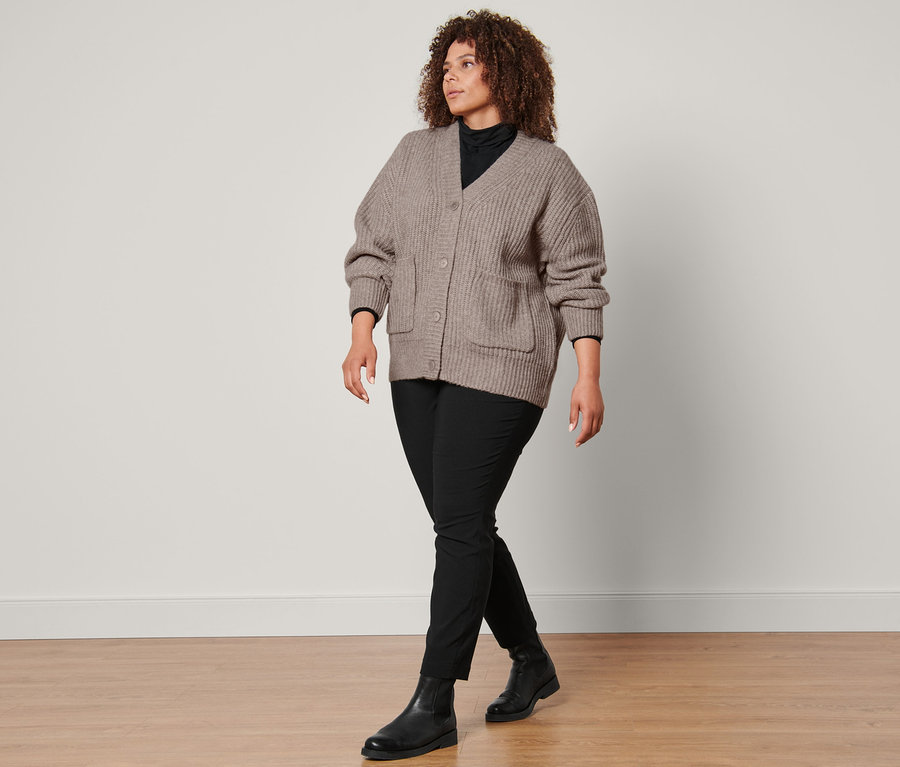 Une femme marche et porte un cardigan en grosse maille avec de la laine, un t-shirt col roulé noir, un pantalon prêt à porter en bengaline noir et des bottines en cuir.