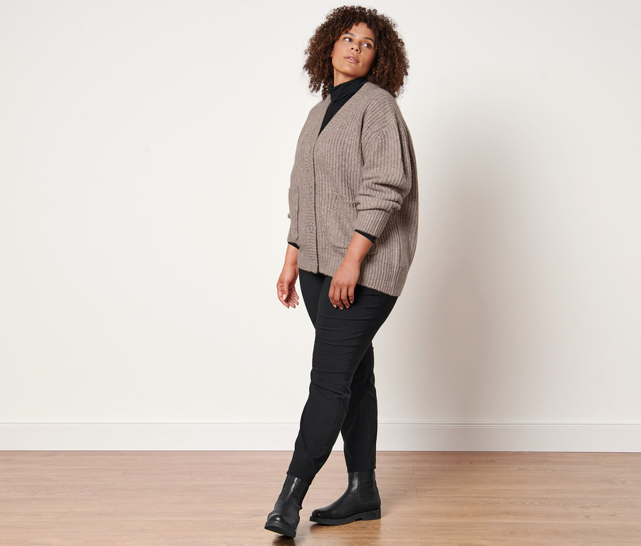 Femme posant en cardigan en grosse maille avec de la laine, un t-shirt col roulé noir, un pantalon en bengaline noir et des bottines en cuir.