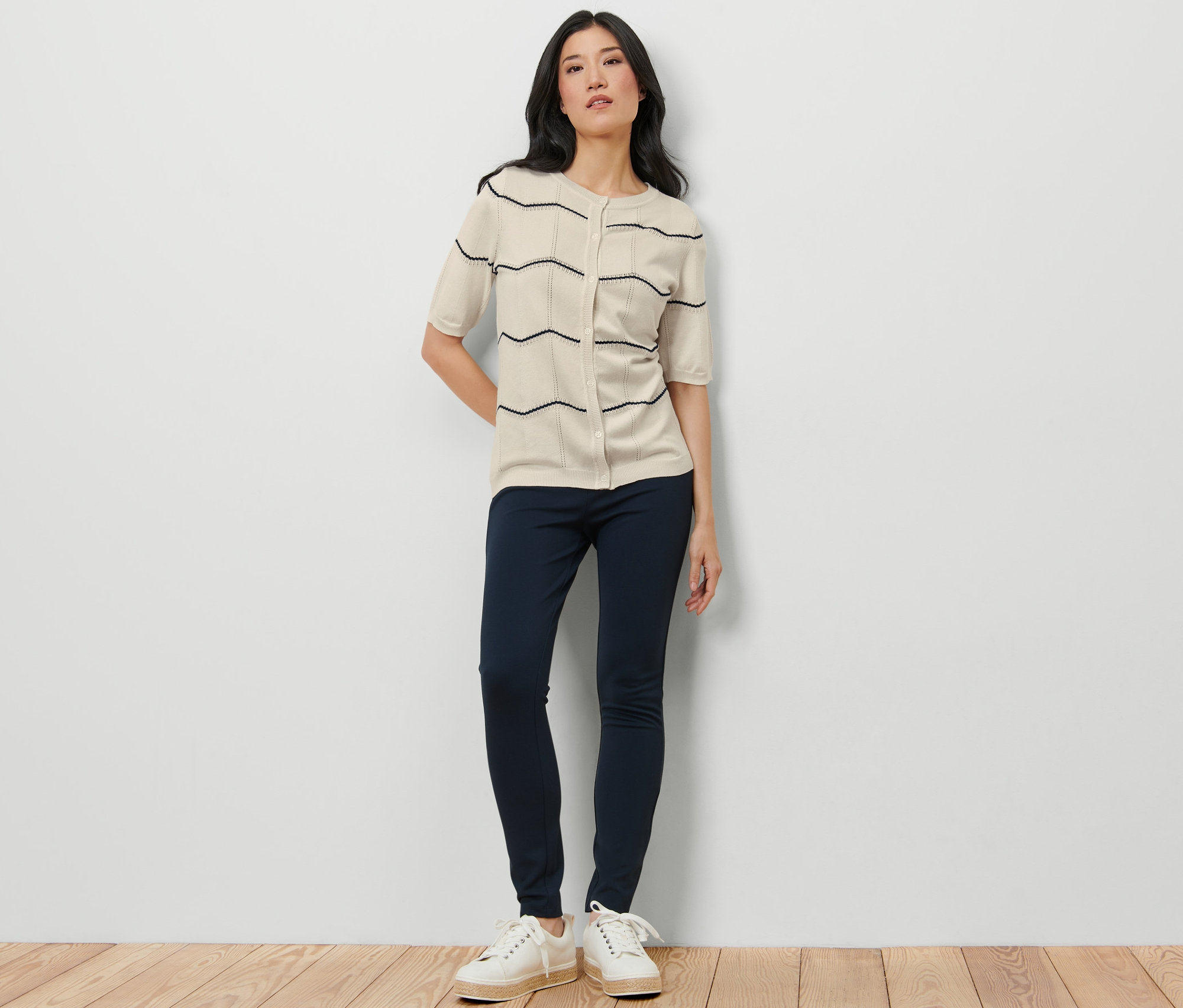 Une femme debout portant un cardigan en maille fine à motif ajouré, un pantalon bleu marine et des baskets blanches.