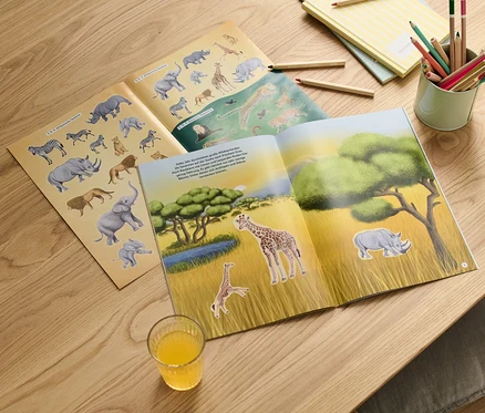 Livre d'autocollants ouvert « Faszierende Stickerwelt – wilde Tiere » avec des images d'animaux sauvages sur une table en bois.