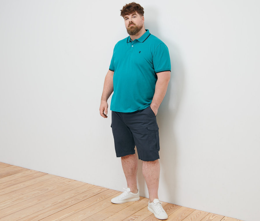 Un homme pose en polo en piqué bleu turquoise et short cargo bleu marine.