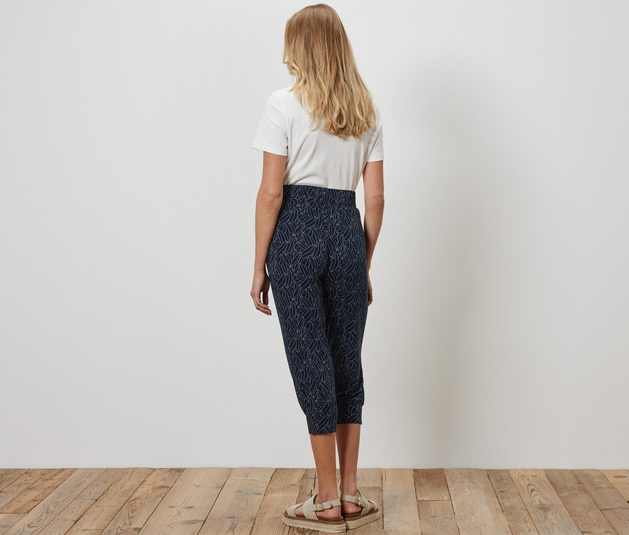 Une femme vue de dos se tient sur un plancher en bois et porte un pantalon 3/4 en jersey bleu-blanc crème et un t-shirt blanc.