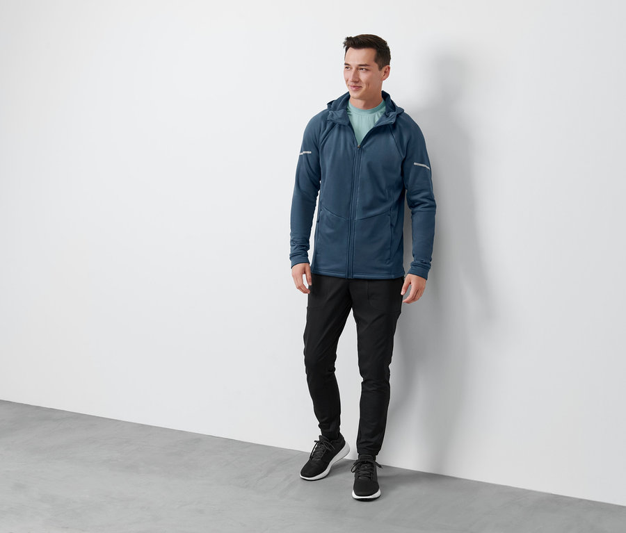 Un homme debout portant une veste thermique bleue, une veste thermique noire, un pantalon fonctionnel thermique et des chaussures noires.