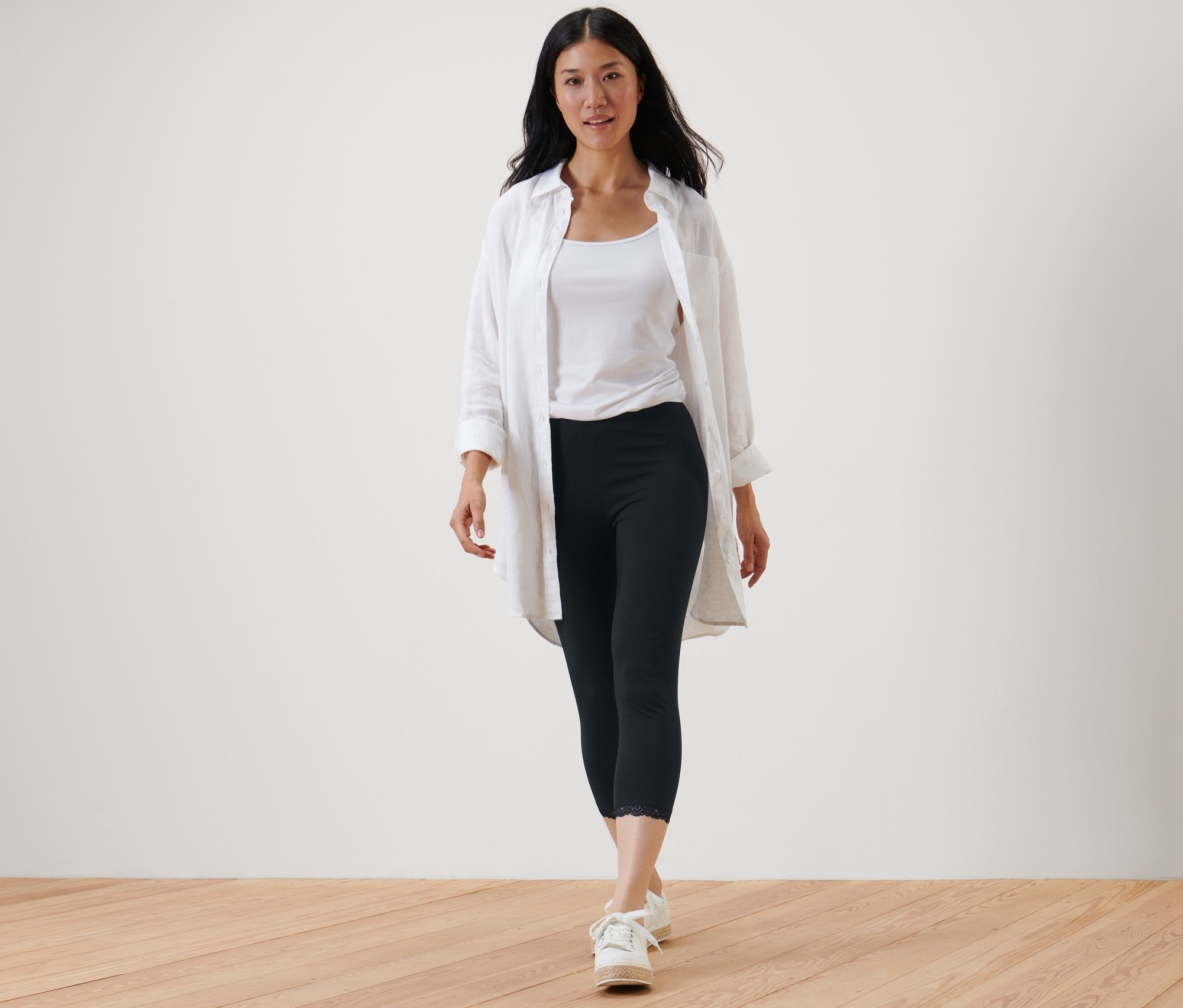 Une femme marche et porte un legging 3/4 avec bord en dentelle noir, un débardeur blanc et une chemise blanche.