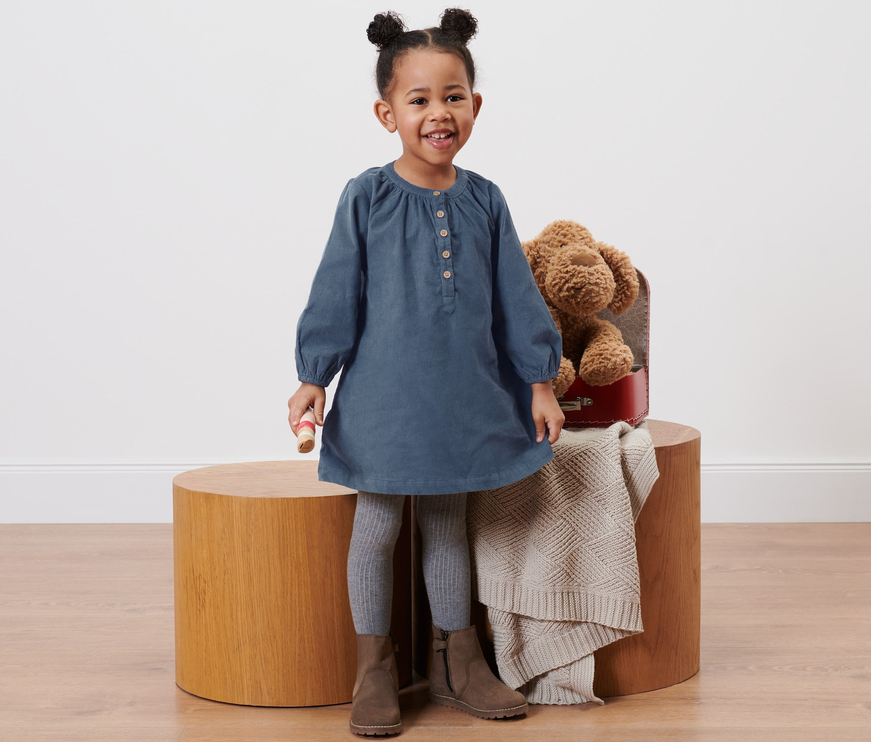 Une petite fille souriante est debout et porte une robe en velours pour enfant, des leggings gris et des bottes marron.
