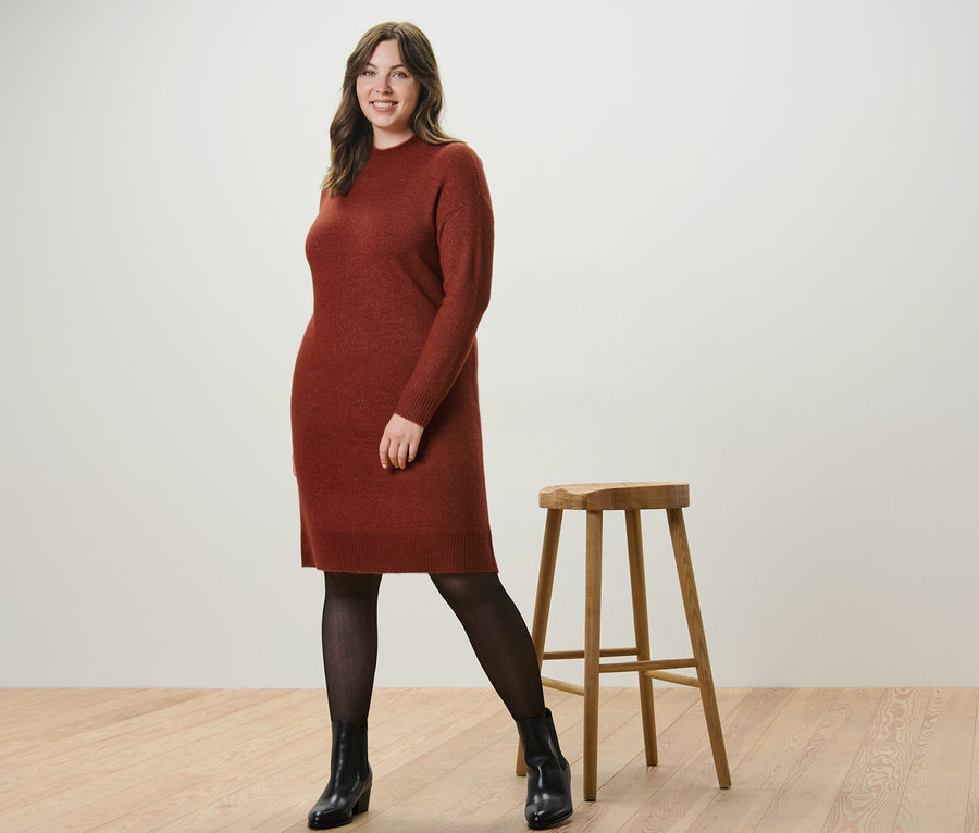 Une femme pose dans une robe en maille avec teneur en laine et des collants fins 50 DEN en aspect transparent à côté d'un tabouret en bois.