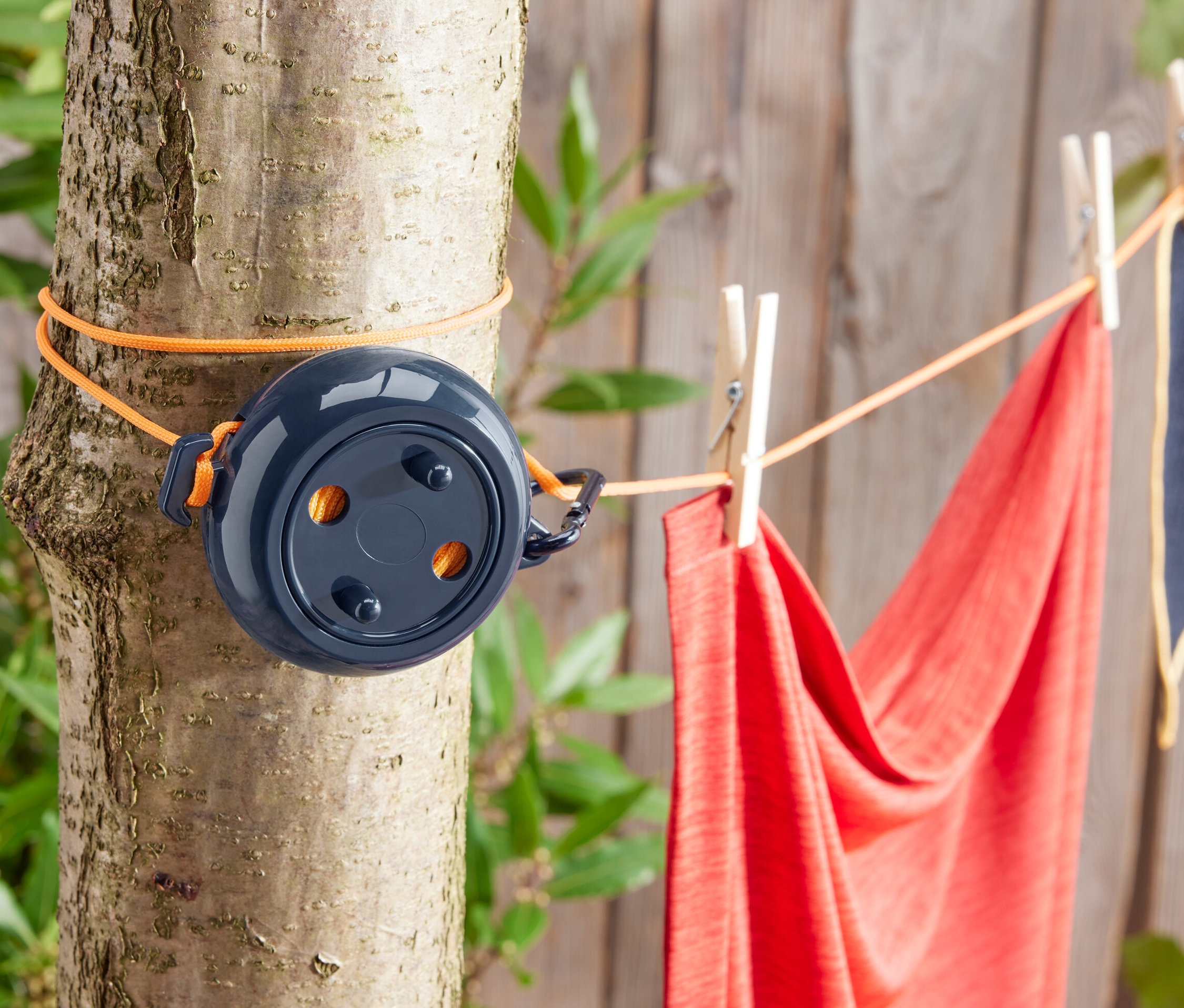 Une corde à linge de camping est fixée à un arbre avec une corde orange. Une serviette rouge et une serviette bleue sont accrochées à la corde à linge avec des pinces à linge.