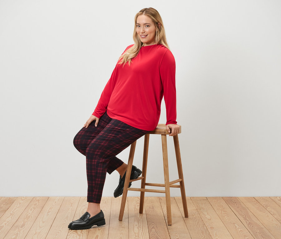Une femme est assise sur un tabouret et porte un pantalon stretch à carreaux et une blouse avec des fronces.